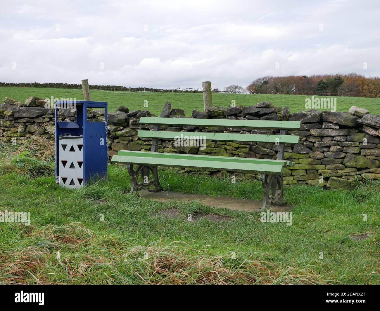 Green and black park bench grey and blue litter bin stood on grass in front of dry stone wall with trees and fields in the background Stock Photo