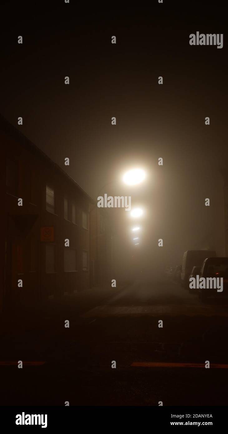 Scary narrow road during a misty autumn night in november during corona pandemic times with street lights like gazing alien eyes Stock Photo