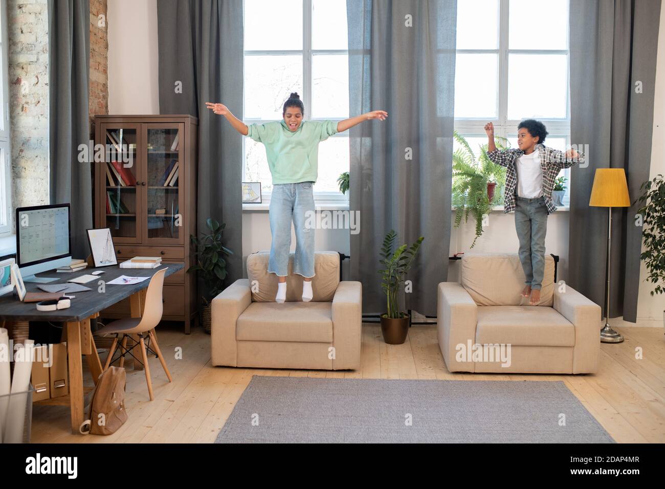 Two cute mixed-race siblings in casualwear jumping on soft armchairs by windows Stock Photo