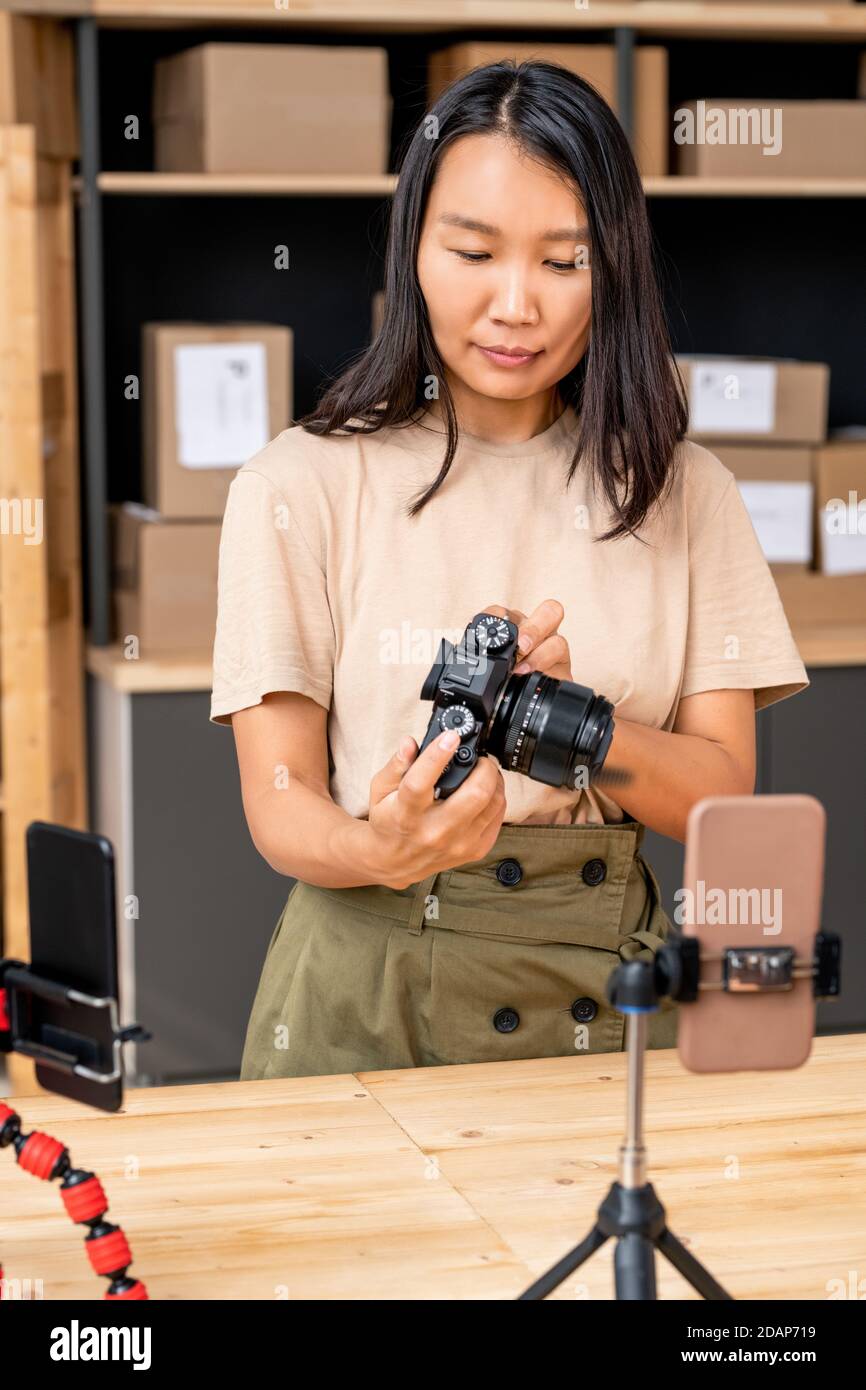 Asian woman in casualwear holding photocamera over table while unpacking it Stock Photo