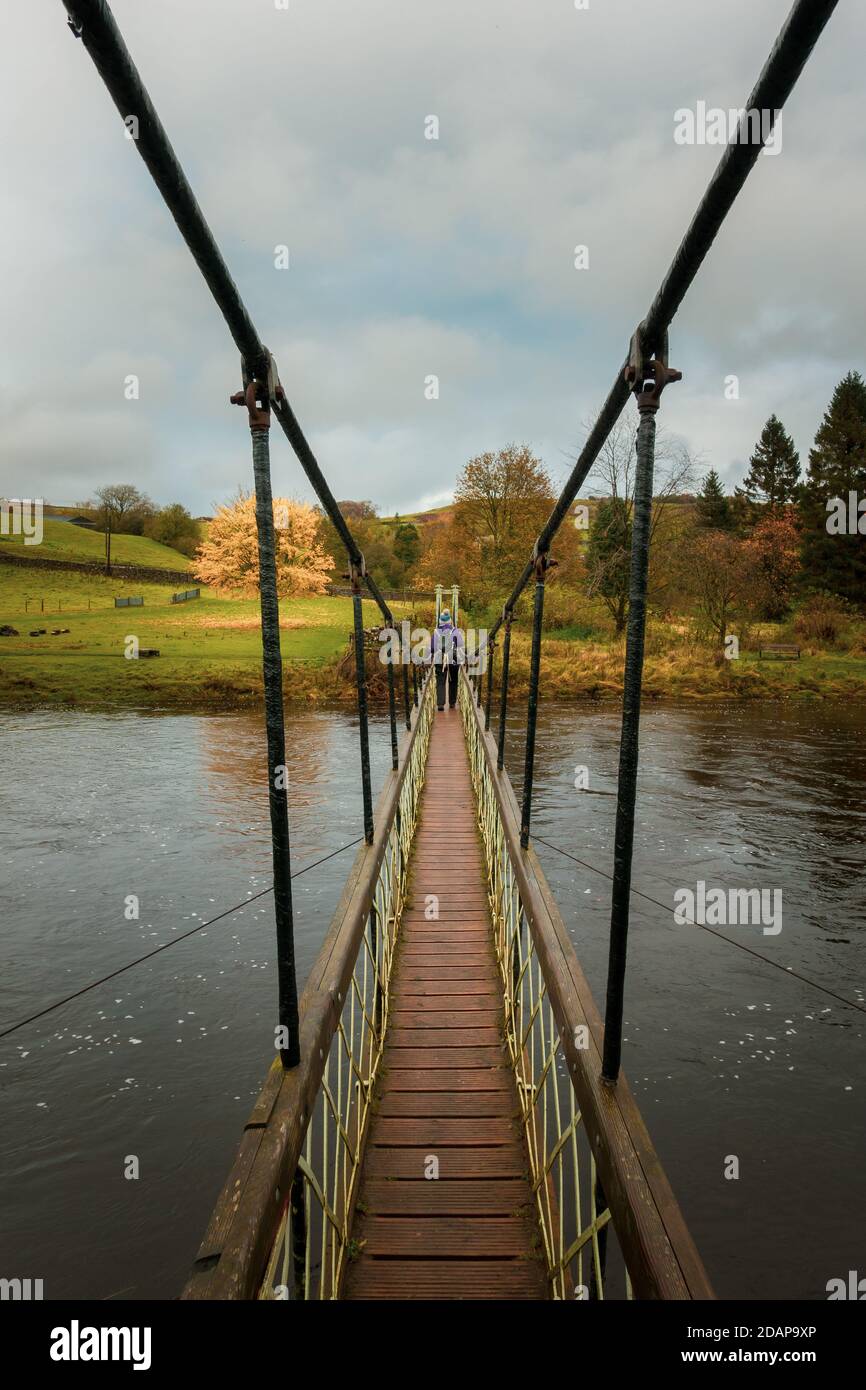 Walker crosses the Hebden suspension bridge on the Dales Way over the River Wharfe in autumn, Yorkshire Dales National Park, UK landscape Stock Photo
