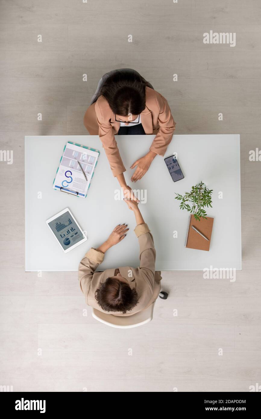 Top view of two elegant businesswomen shaking hands over desk after negotiating Stock Photo