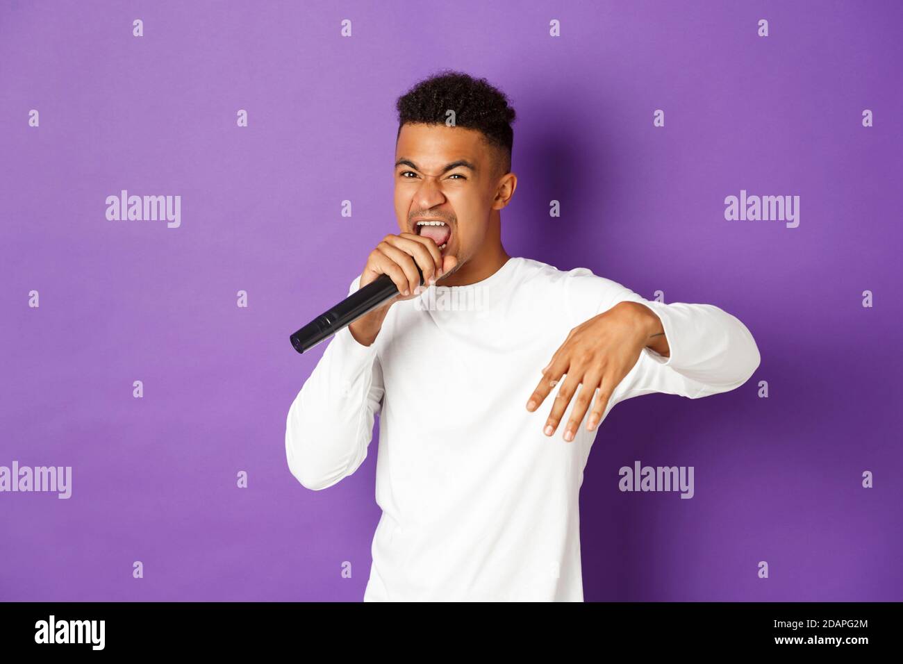 Image of sassy african-american guy singing in microphone, rapping and performing on stage, standing over purple background Stock Photo