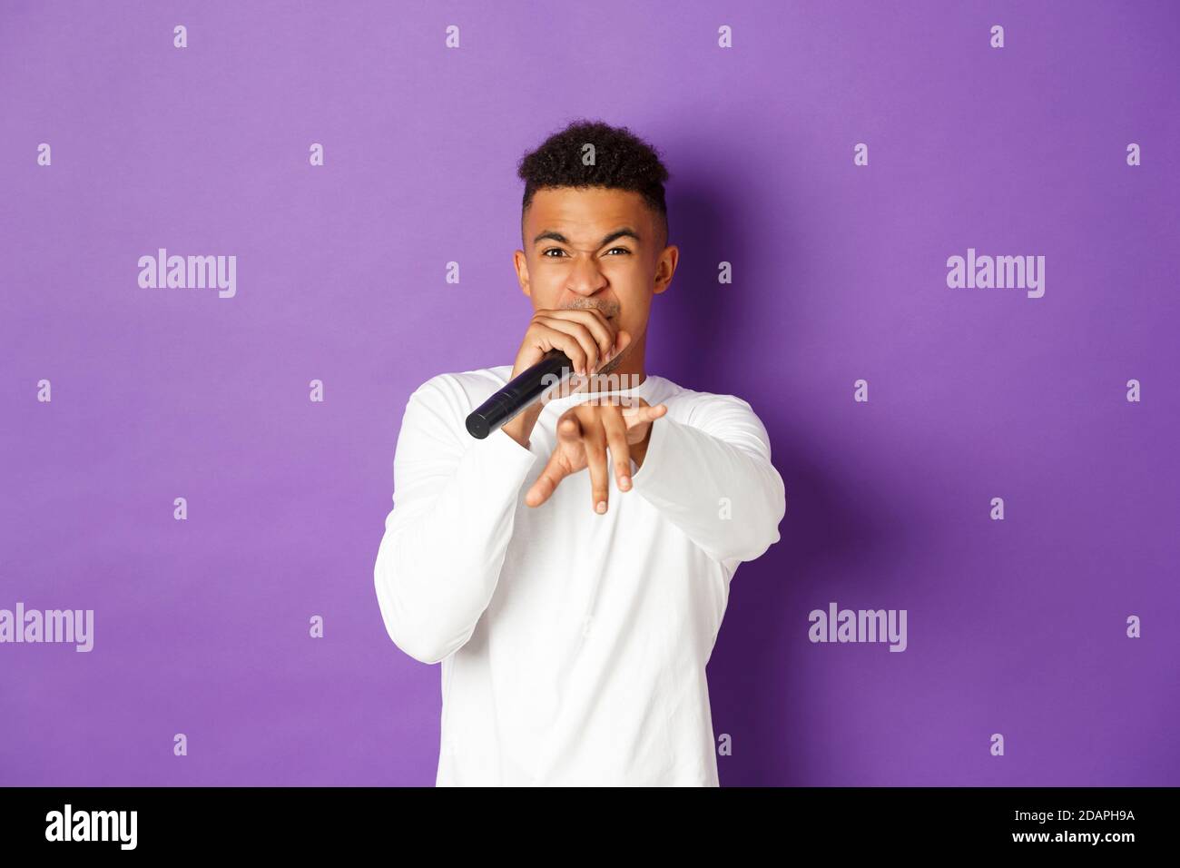 Image of sassy african-american guy singing in microphone, rapping and pointing at camera, standing over purple background Stock Photo