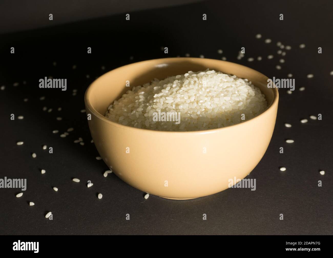 White uncooked rice in a bowl with scattered grains nearby on a black background Stock Photo