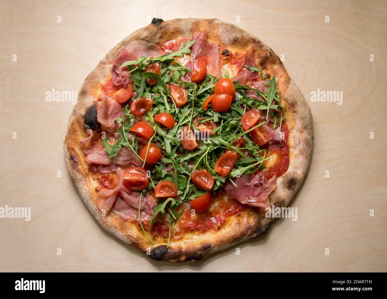 Italian pizza topped with tomato sauce, mozzarella, prosciutto, rocket salad and cherry tomatoes, top view on wooden background Stock Photo
