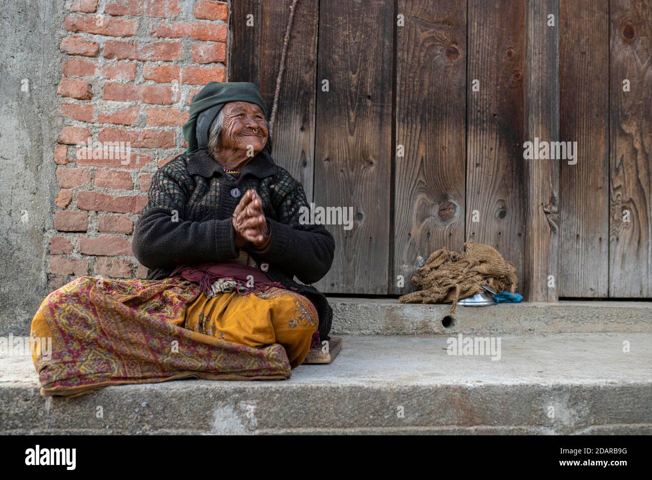 Friendly smiling elderly woman with headscarf and nose ring sitting in front of a wooden door, Himalaya, Jiri, Khumbu region, Nepal Stock Photo