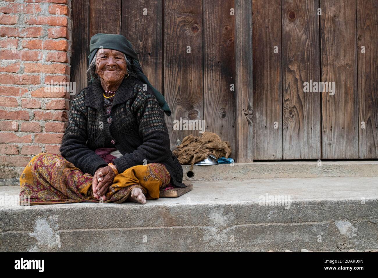 Friendly smiling elderly woman with headscarf and nose ring sitting in front of a wooden door, Himalaya, Jiri, Khumbu region, Nepal Stock Photo