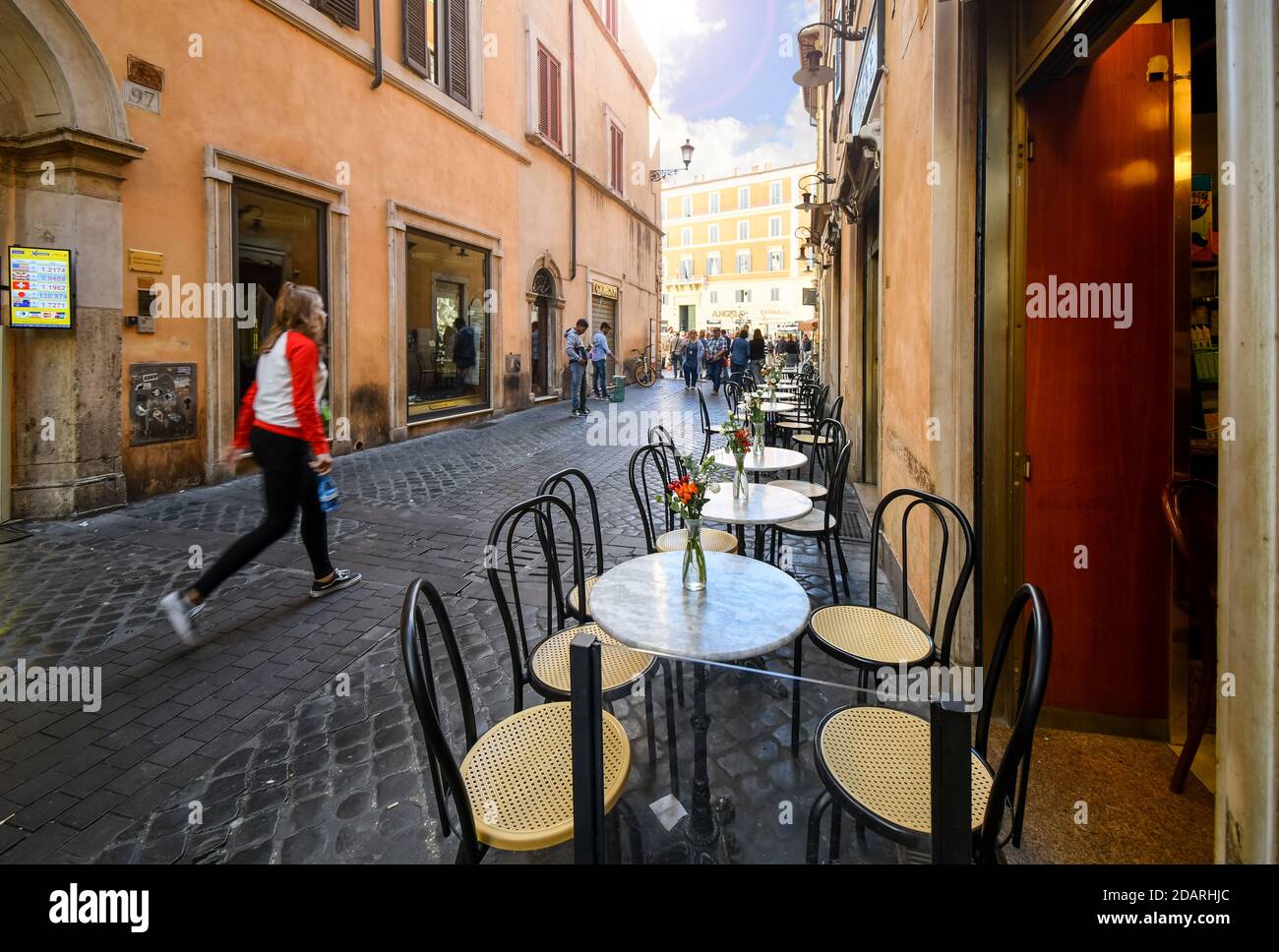 Tourists walk down a narrow cobbled street lined with patio outdoor tables from a cafe in the center of Rome, Italy Stock Photo