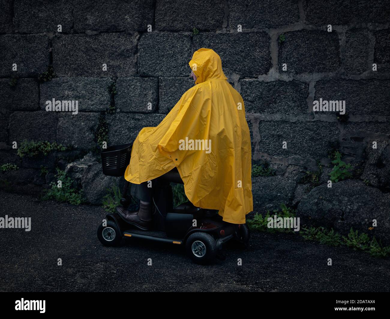 Man on a Mobility Scooter / Vehicle wearing a Yellow Raincoat / Poncho / Cagoule / Jacket, in profile with Old Grey Stone Wall Background Stock Photo