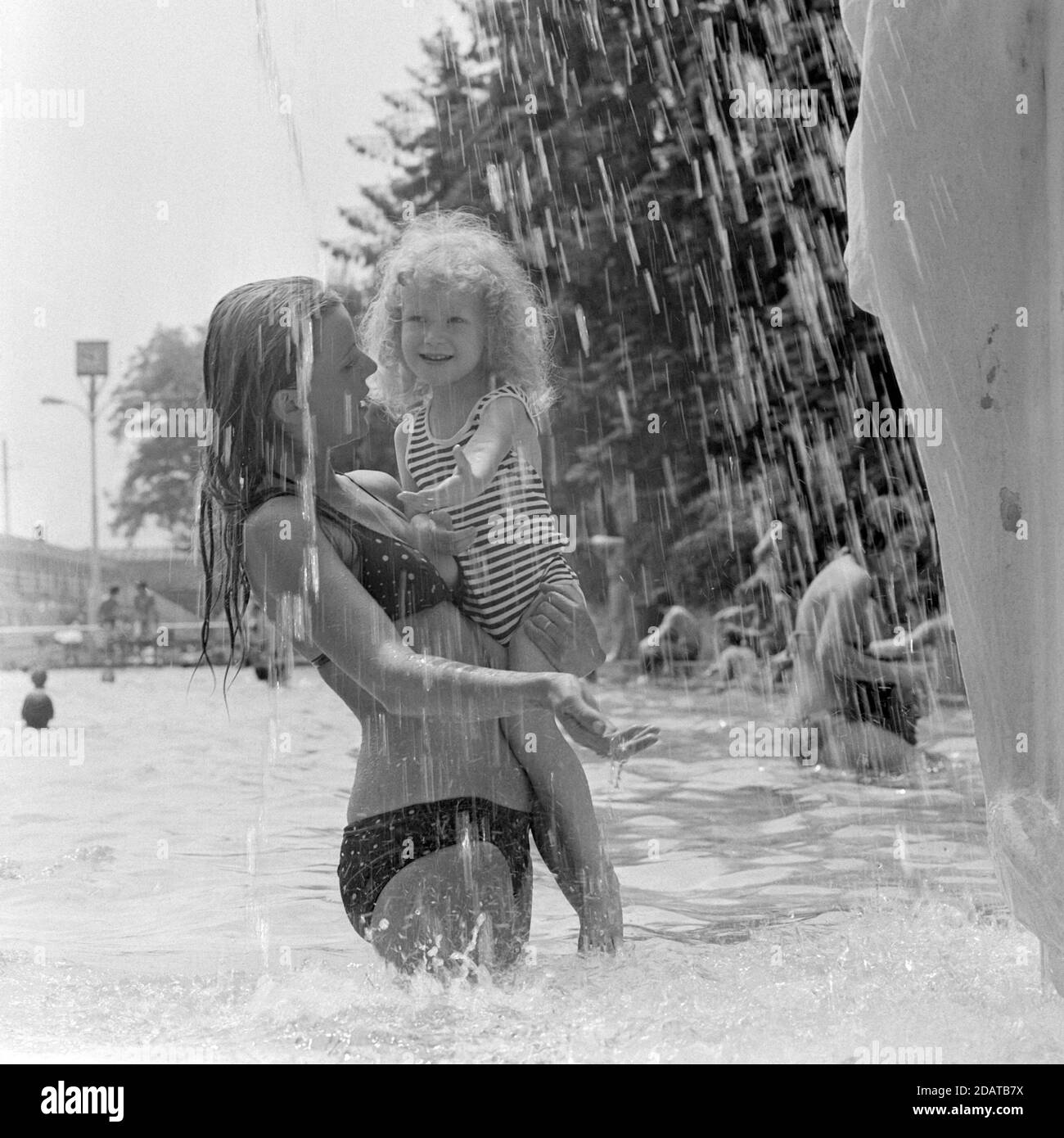 mother holding young daughter beneath fountain in public swimming pool 1970s hungary Stock Photo