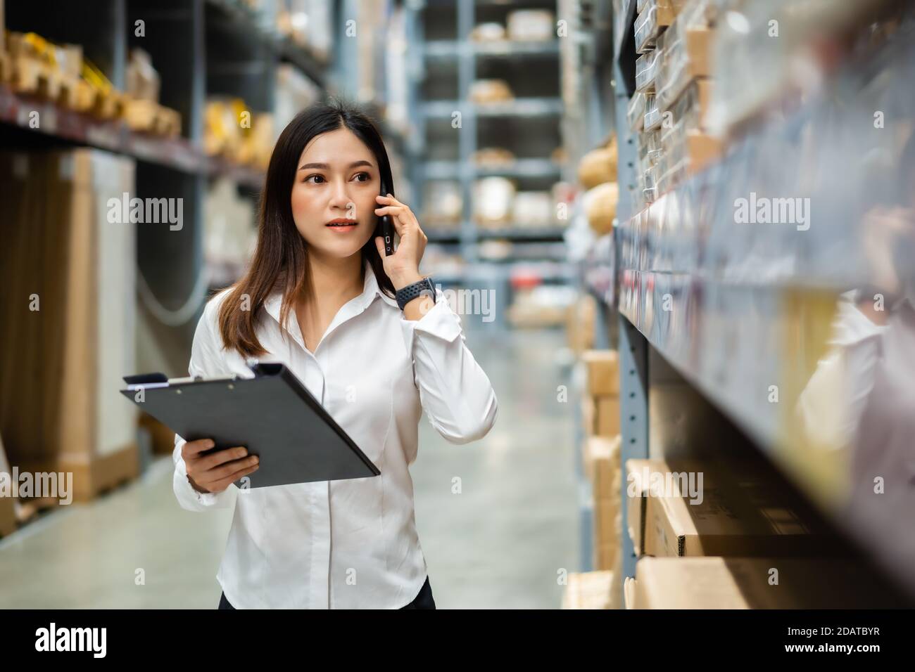 young woman worker talking on a mobile phone and holding clipboard to checking inventory in the warehouse store Stock Photo