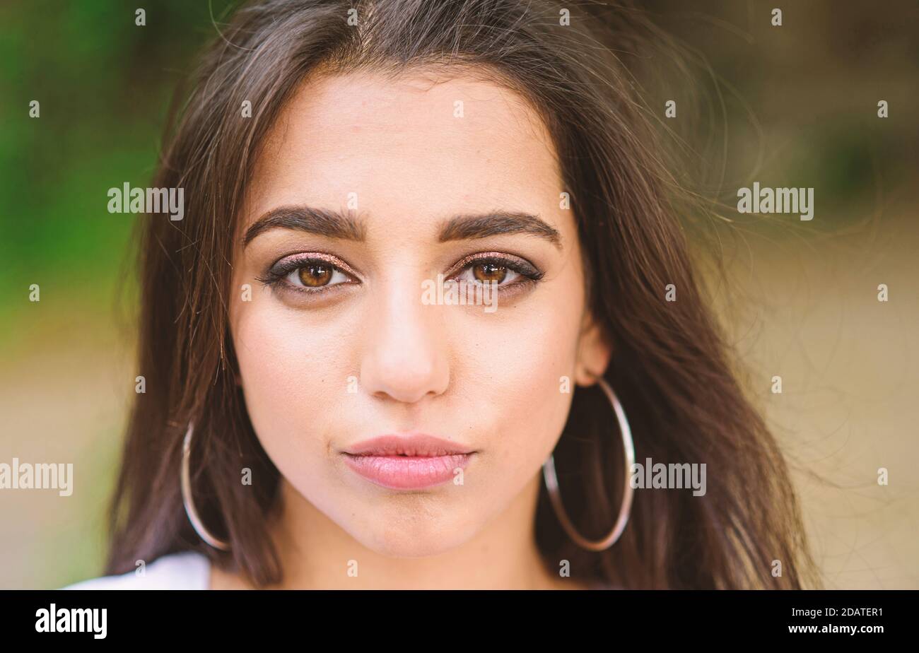 Girl attractive gorgeous brunette middle eastern appearance close up defocused background. Beauty of arabian women. Girl wears big metallic ring earrings. Woman confident face make up looks at camera. Stock Photo