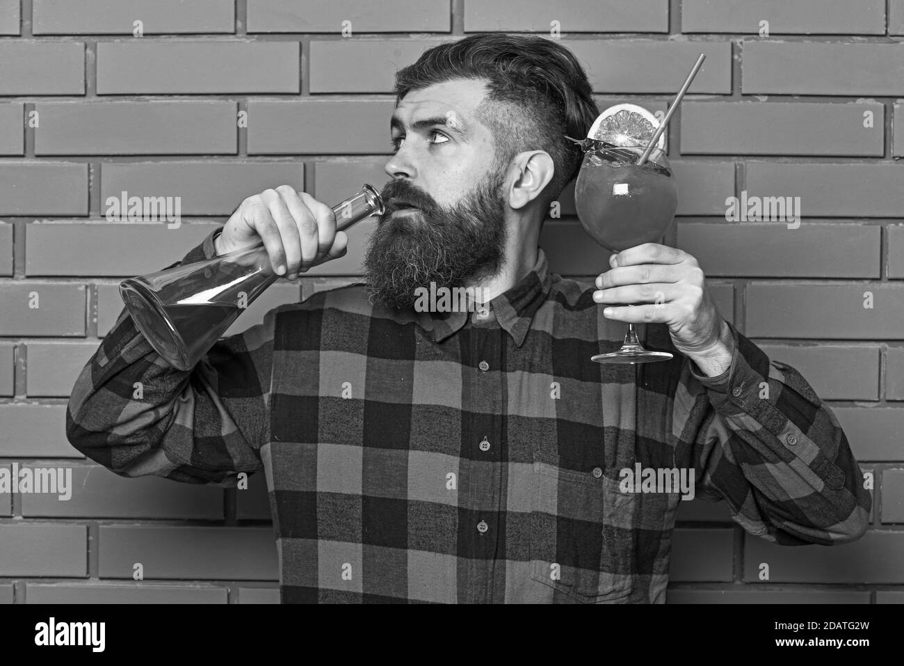 Barman with beard and strict face drinks out of bottle with alcohol. Man holds glass and bottle on brick wall background. Barman or hipster holds drink with orange and straw. Drinking alcohol concept. Stock Photo
