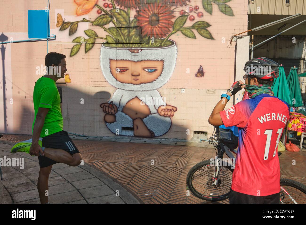A jogger & a cyclist chatting in front of a mural by artist Alex Face featuring his character Mardi meditating; Phahurat / Chinatown Bangkok, Thailand Stock Photo
