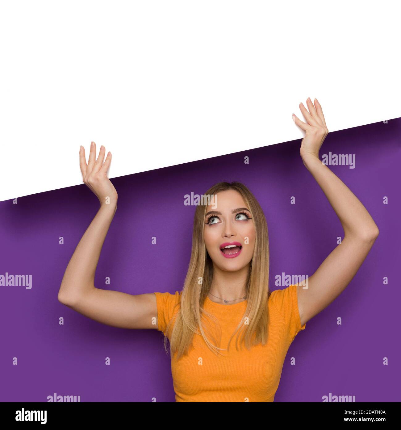 Beautiful young woman in orange top is holding white banner over head, looking up and talking. Front view. Waist up studio shot on purple background. Stock Photo