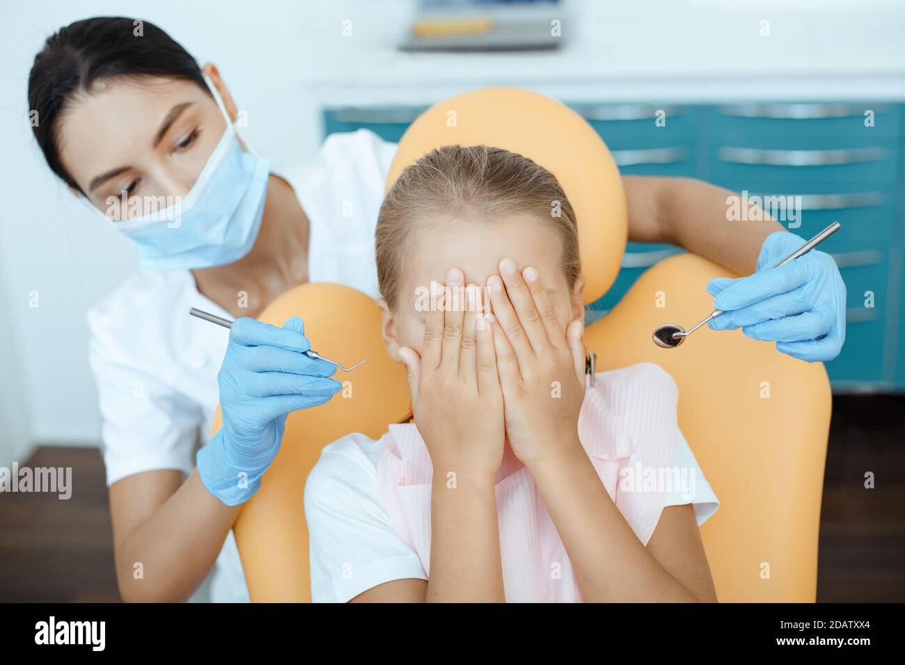 Little child covers face with hands, sits in medical chair with woman doctor holds instruments for examining patient Stock Photo