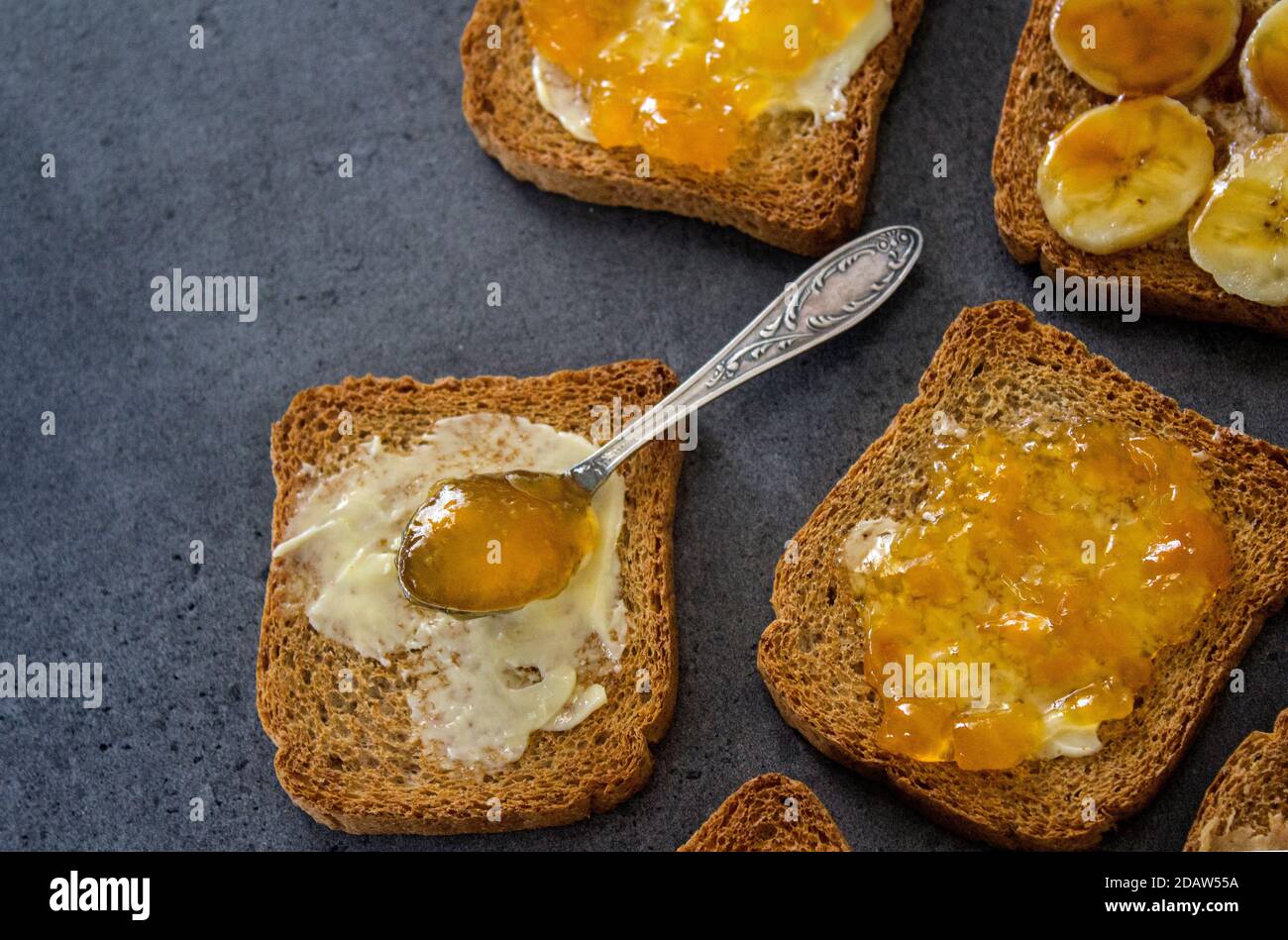 Sweet breakfast top view photo. Toasts with different toppings: peanut butter, jam, butter and caramelized banana. Dark grey textured background with Stock Photo