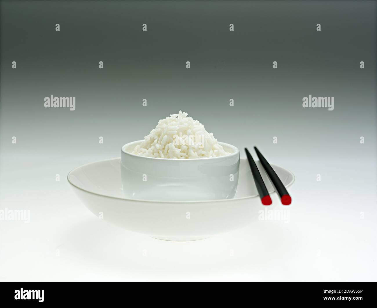A white bowl on a plate, contains rice, with Chinese sticks, on a soft white background, with shadows. Stock Photo
