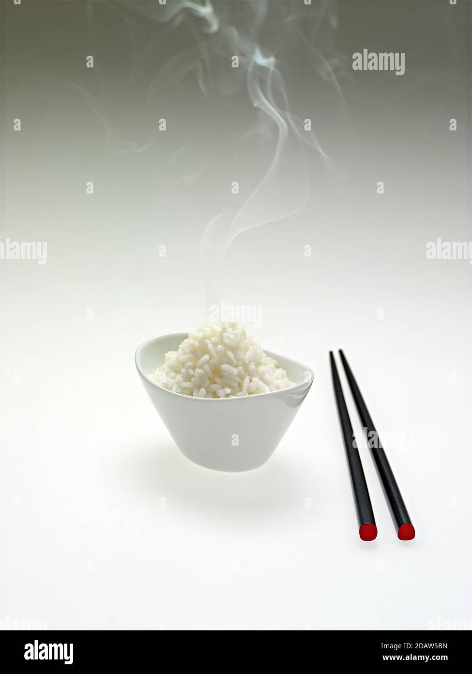 A white bowl contains steaming rice, with Chinese sticks, on a white background, with shadows. Stock Photo