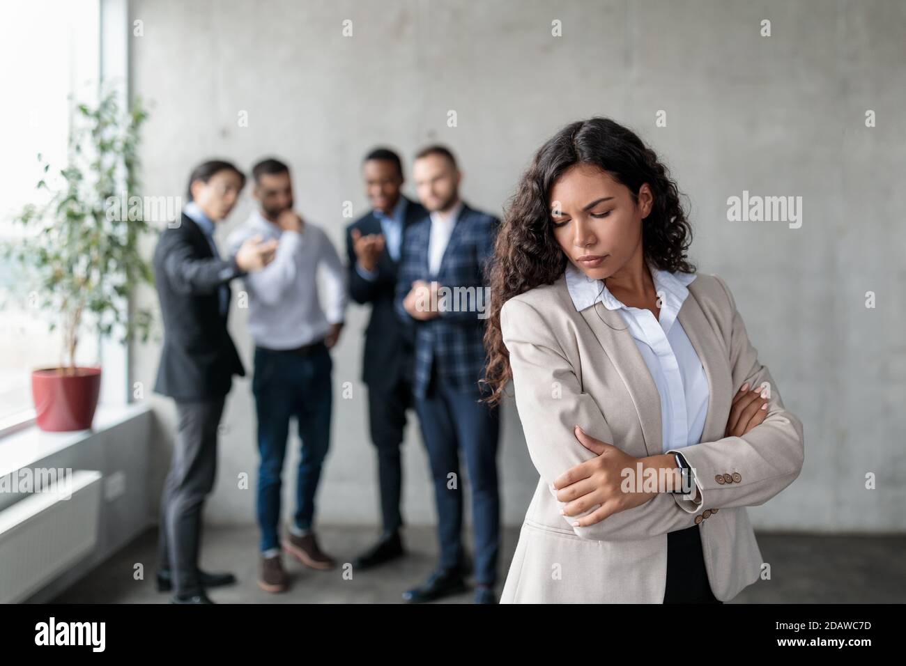 Unhappy Businesswoman Standing While Colleagues Whispering Behind Back In Office Stock Photo