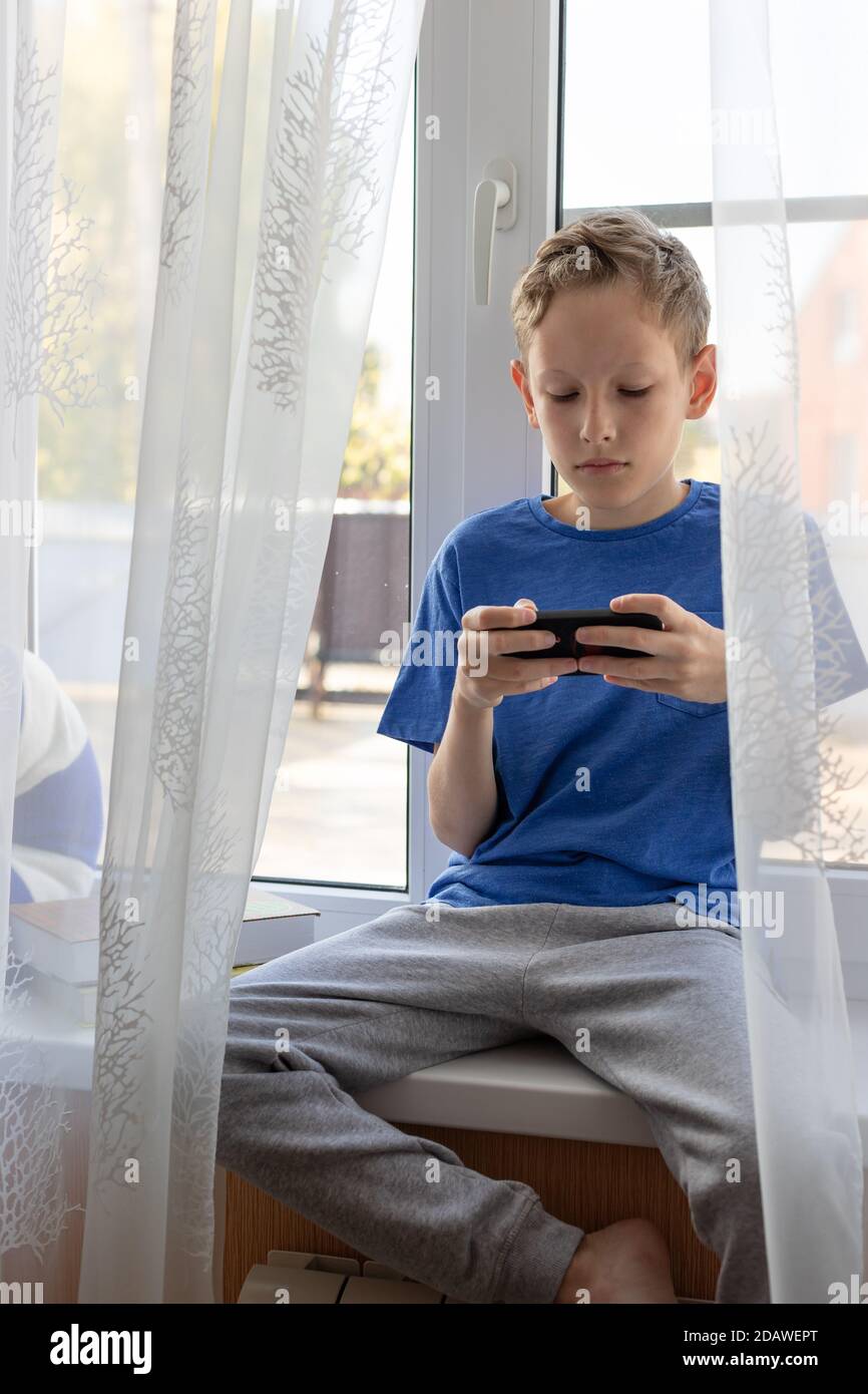 Preteen boy uses mobile phone while sitting on the windowsill at home. Stock Photo