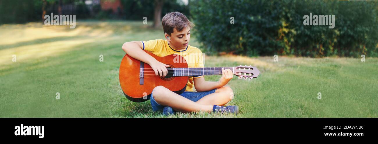 Hard of hearing preteen boy playing guitar outdoor. Child with hearing aids in ears playing music and singing song in park. Hobby art activity Stock Photo