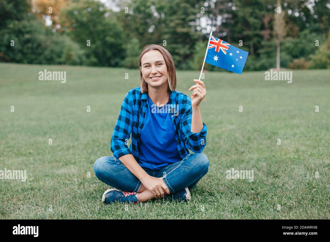 Happy Caucasian middle age woman waving Australian flag. Smiling proud citizen sitting on grass in park celebrating Australia Day holiday in January o Stock Photo