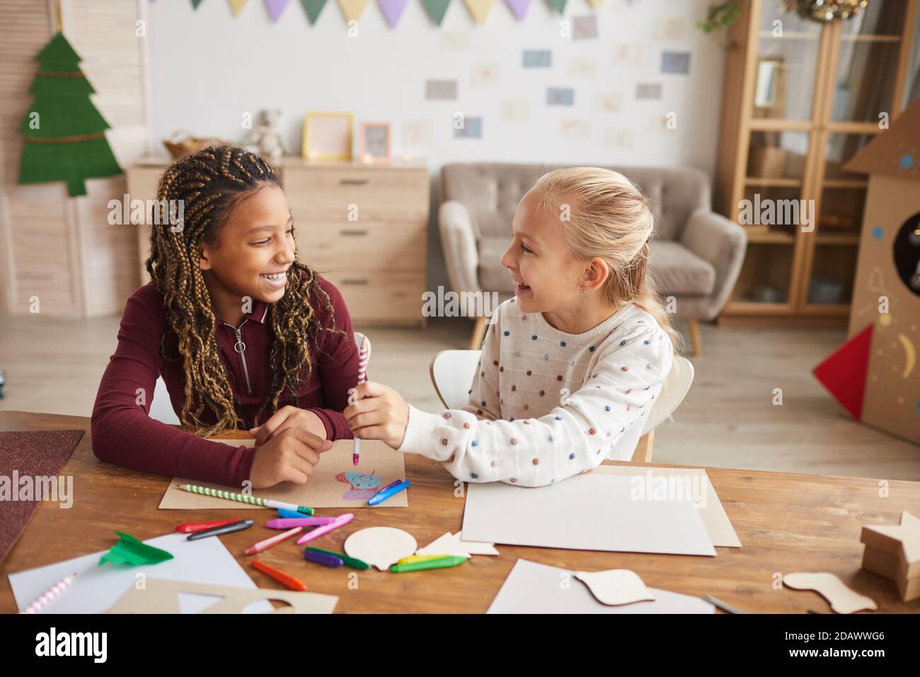 Portrait of two laughing teenage girls enjoying crafting and painting toether while siting at desk in decorated playroom, copy space Stock Photo