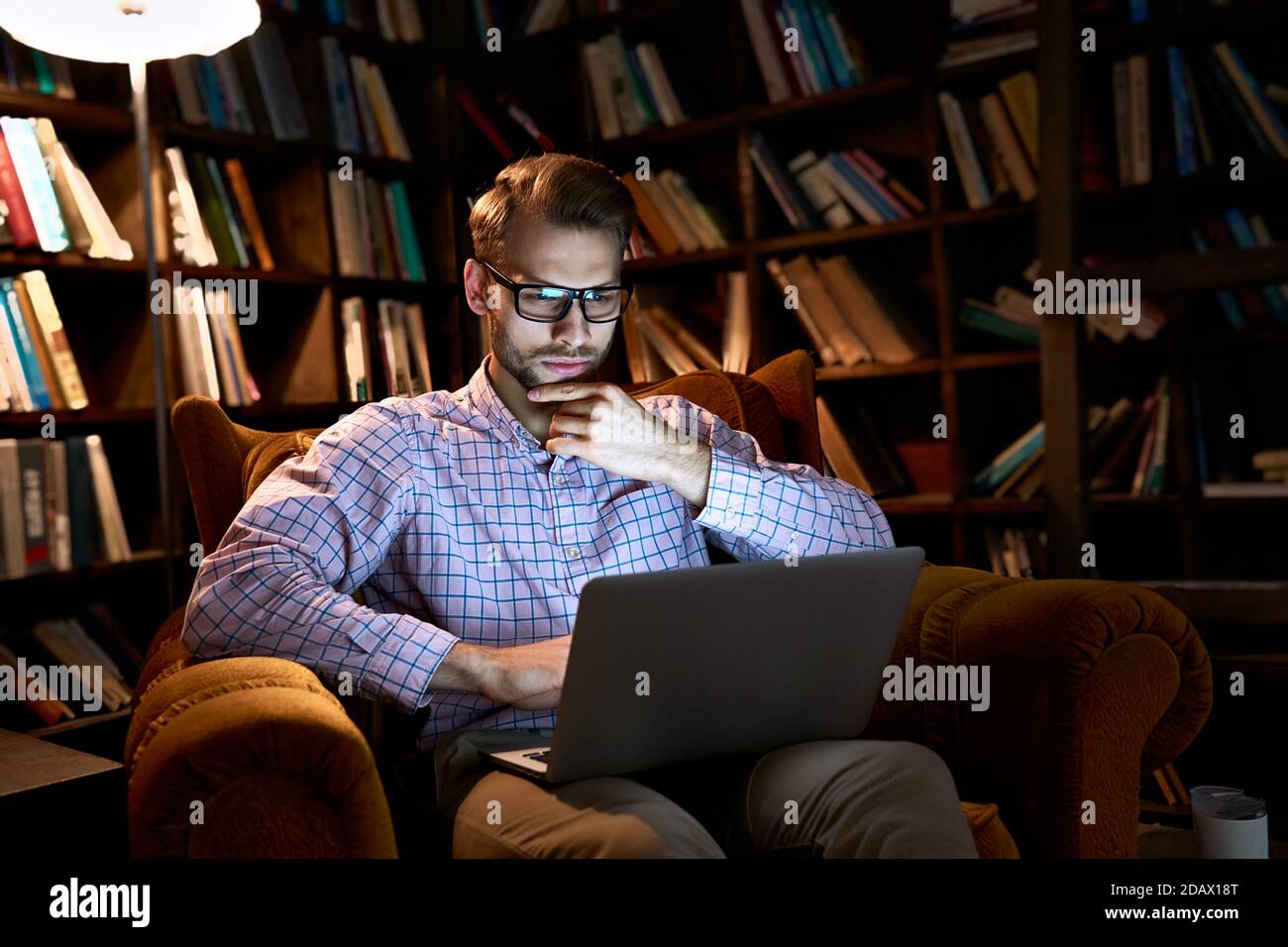 Serious young business man using laptop working late online sitting in chair. Stock Photo