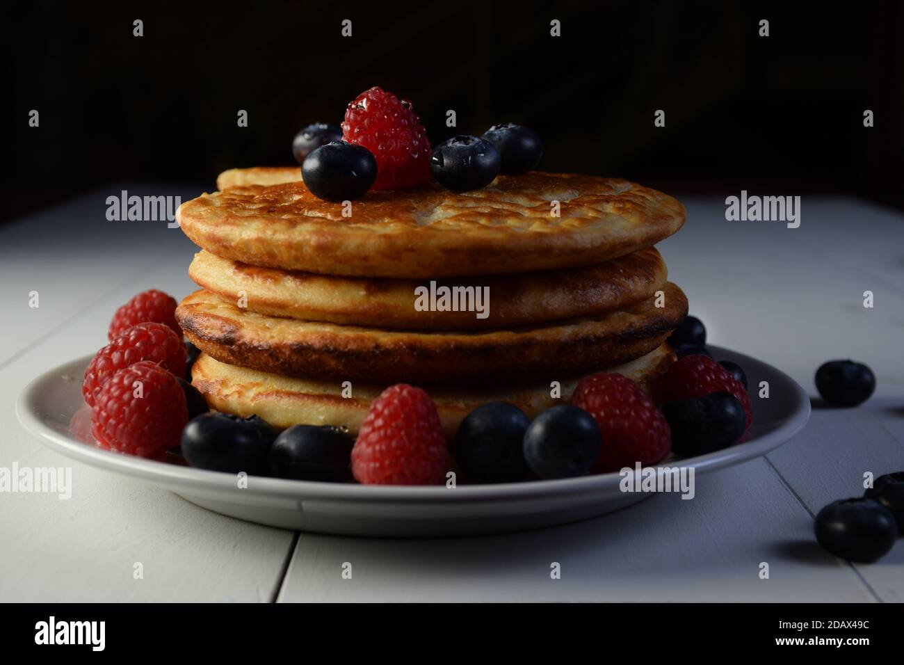 Plate of pancakes with maple syrup, blueberries and raspberries on white wooden table and black background. Stock Photo