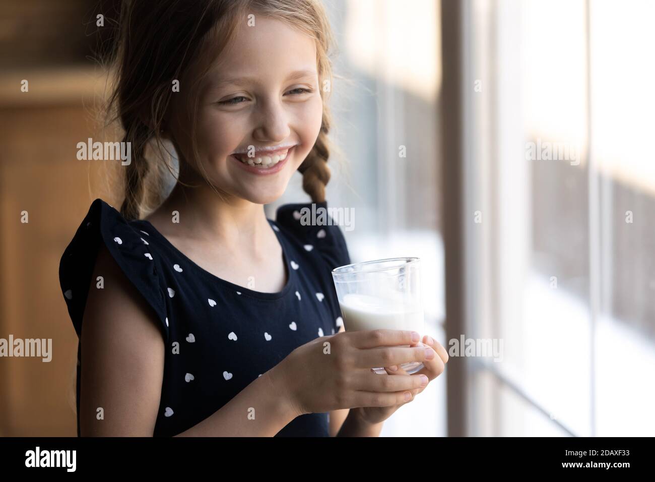 Naughty little girl standing by window drinking dairy from glass Stock Photo