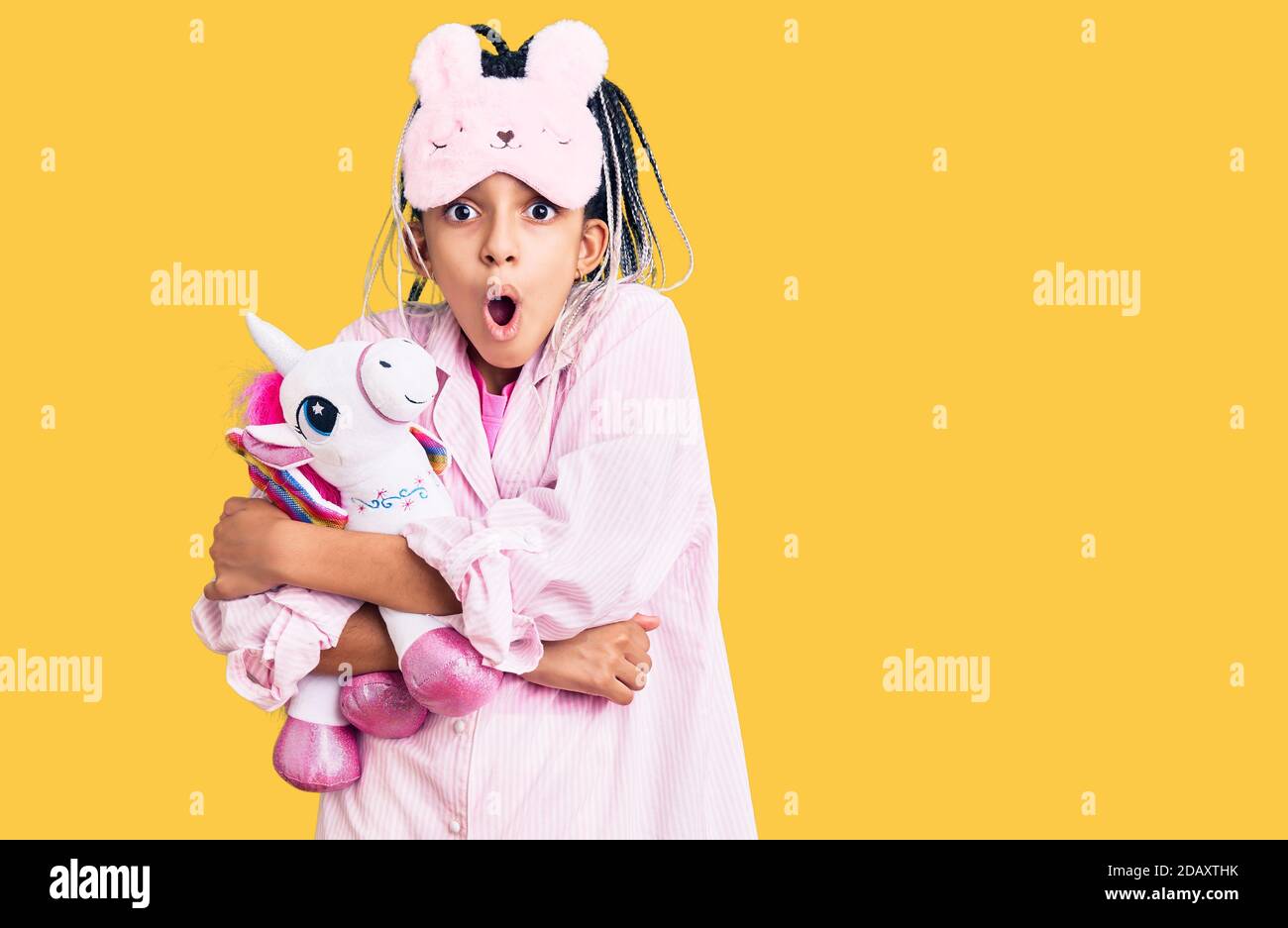 Cute african american girl wearing sleep mask and pajama holding pink teddy unicorn scared and amazed with open mouth for surprise, disbelief face Stock Photo