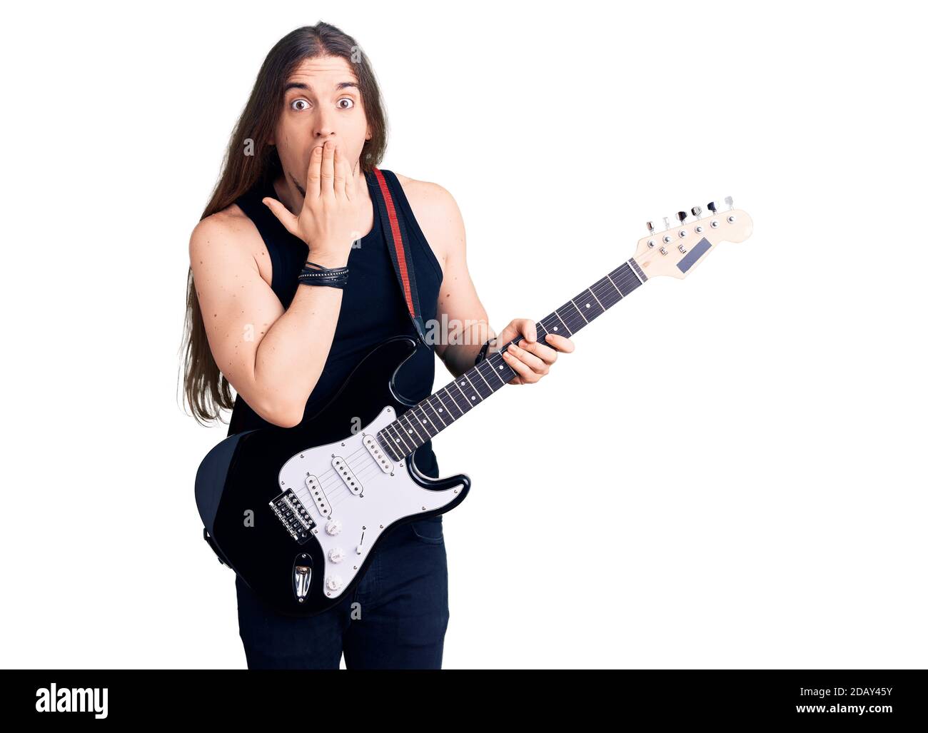 Young adult man with long hair playing electric guitar covering mouth with hand, shocked and