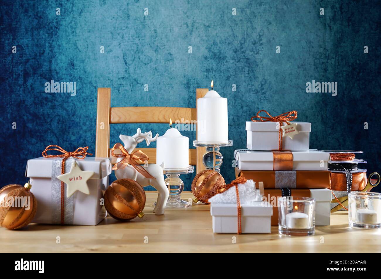 Festive Christmas gifts and gift wrapping in copper and white theme, with candles and ornaments on a natural wood table against a blue background. Neg Stock Photo