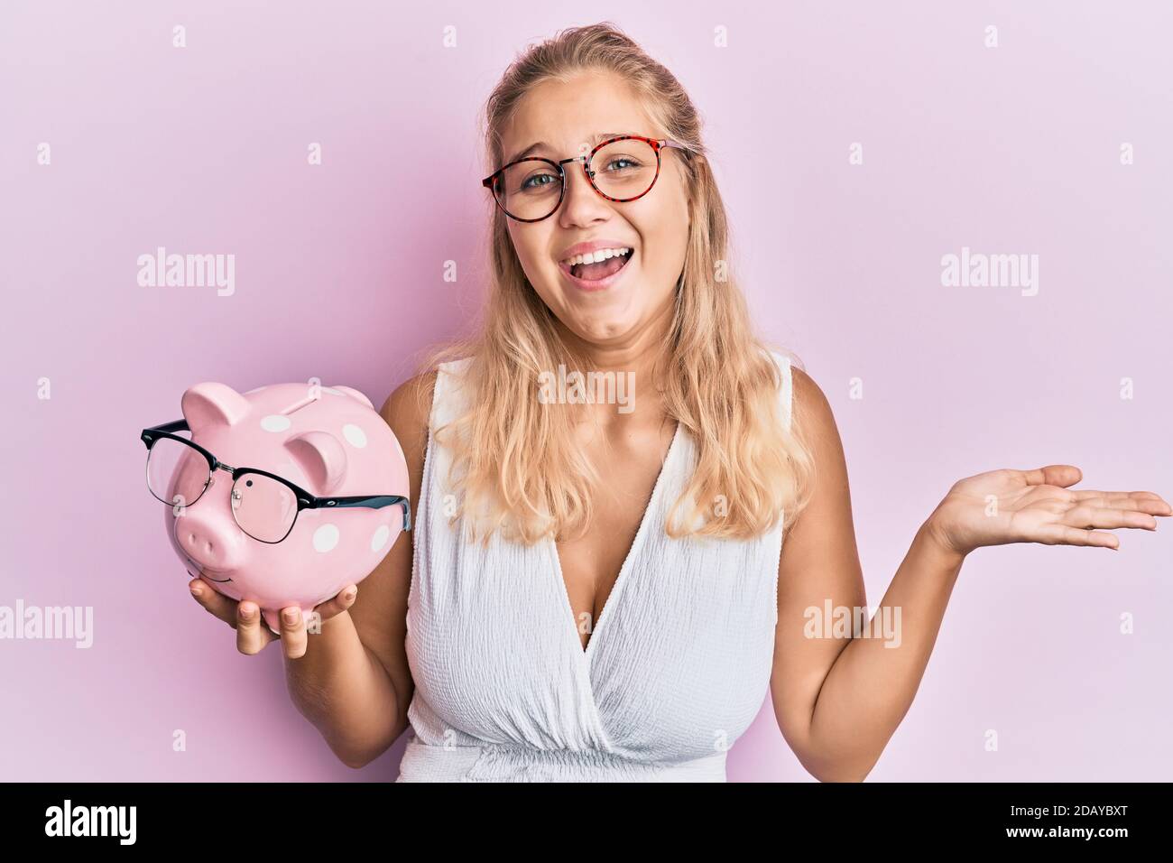 Young blonde girl holding piggy bank with glasses celebrating victory ...