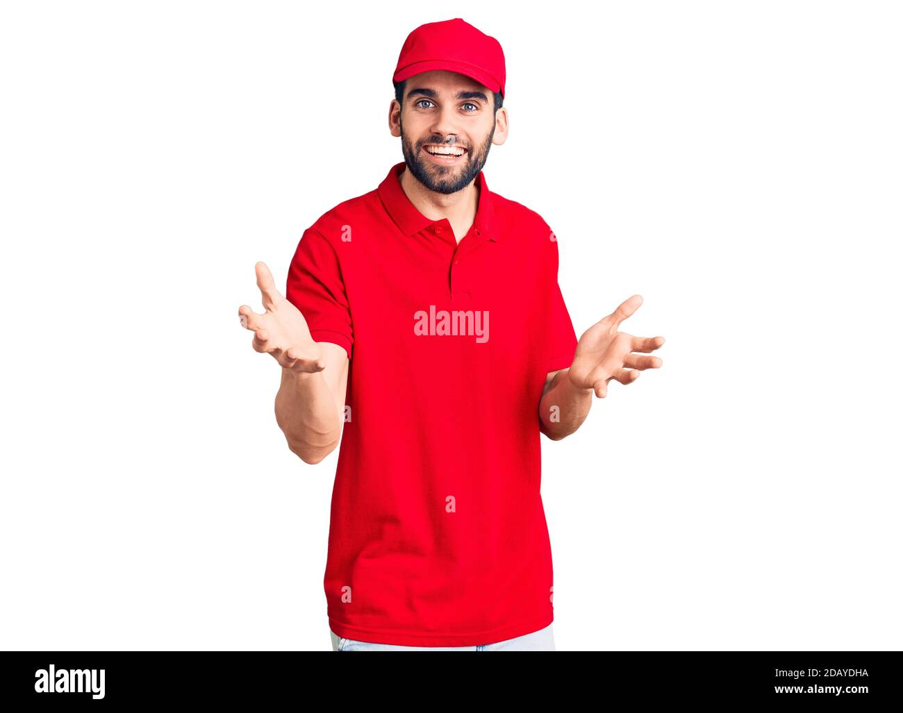 Young handsome man with beard wearing delivery uniform smiling cheerful with open arms as friendly welcome, positive and confident greetings Stock Photo