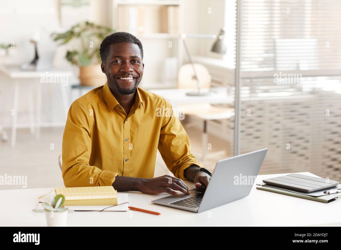 Portrait of smiling African-American man using laptop and looking at camera while enjoying work in minimal office interior, copy space Stock Photo