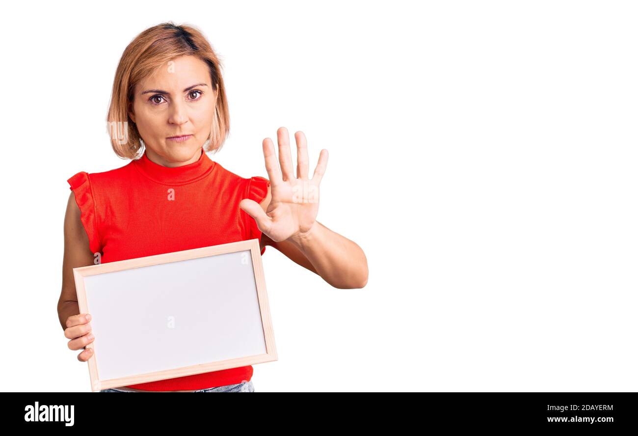 Young blonde woman holding empty white chalkboard with open hand doing stop sign with serious and confident expression, defense gesture Stock Photo