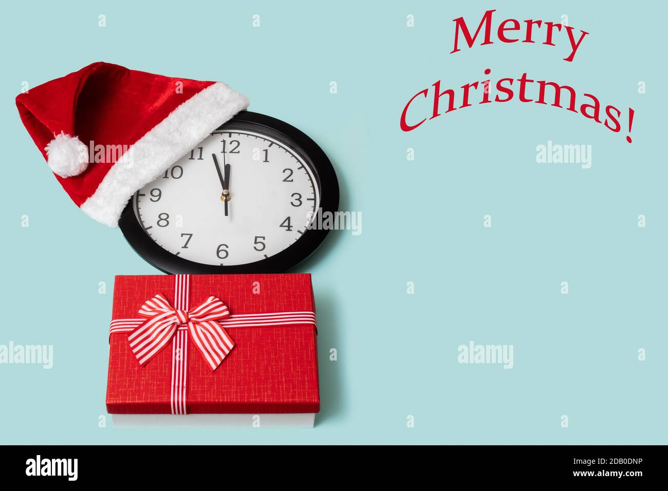 Christmas composition with a classic analog clock in a red Christmas cap and a red gift box with a bow isolated on a blue background with the text Mer Stock Photo