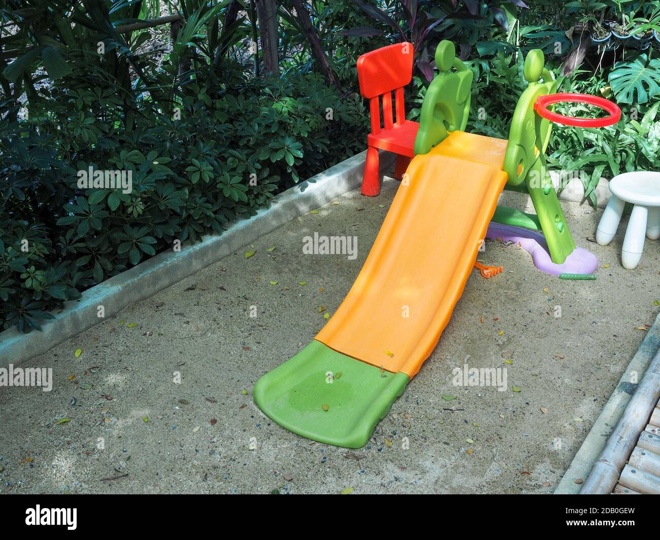 Old empty orange and green color playground slide for kid on sand near the garden. Stock Photo