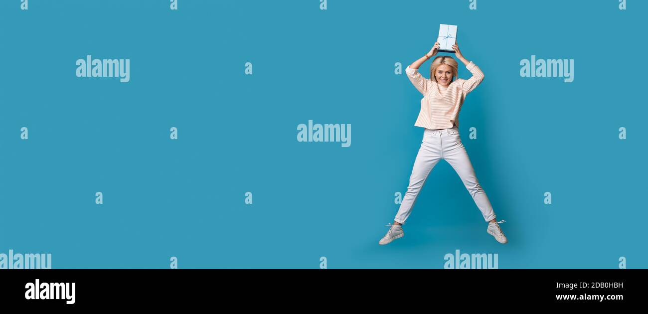 Blonde caucasian woman is jumping on a blue studio wall with free space holding a present box on her head and smile Stock Photo