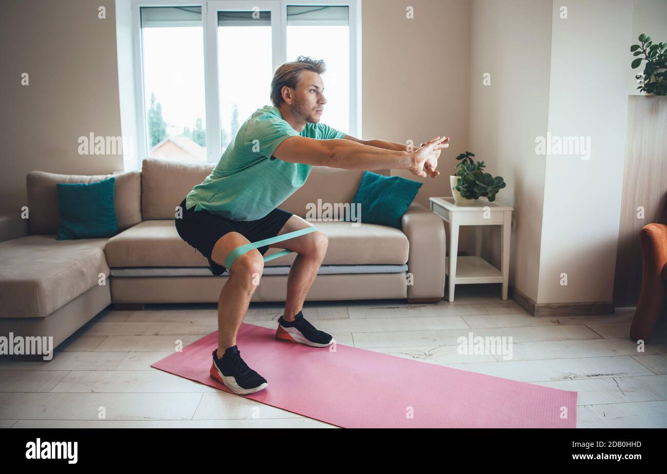 Caucasian man is doing fitness at home using special bands on the floor while squatting Stock Photo