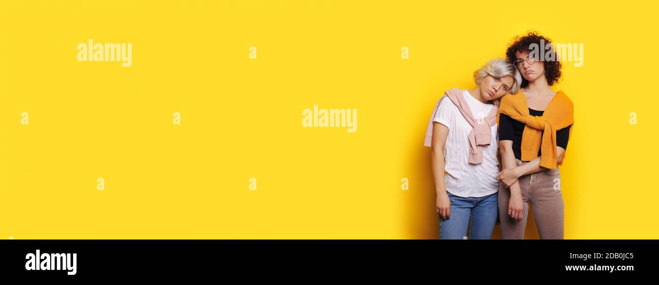 Charming blonde lady lying her head on sisters shoulders feeling sad and posing on a yellow studio wall with free space Stock Photo
