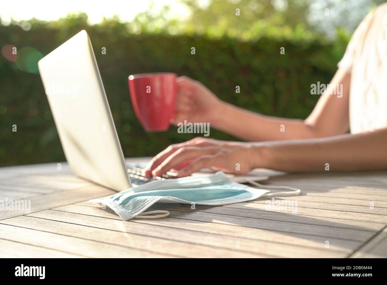 Medical face mask with woman typing and holding a red cup at the background. Work at home concept. Stock Photo