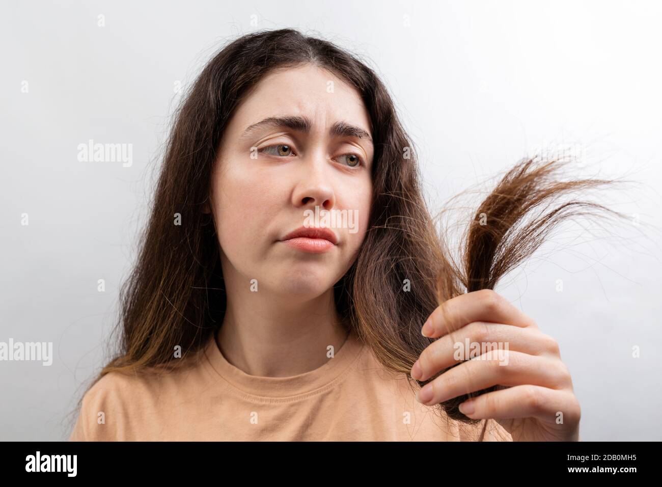 A. young Caucasian woman in a beige t-shirt looks distressed at the dry ends of her long hair. White background. Stock Photo