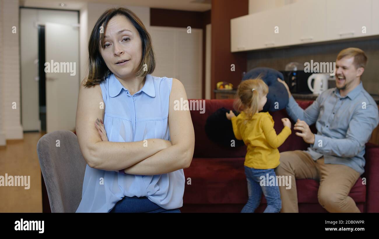 Tired depressed young caucasian mother woman feel very annoyed, suffering migraine of active father and daughter child. Naughty girl jumps, plays noisily with man at home. Hyperractive children Stock Photo