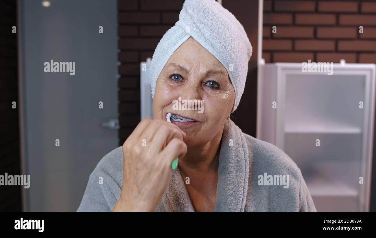 Attractive old senior woman grandmother with towel brushing teeth and  looking into a mirror. Elderly grandma