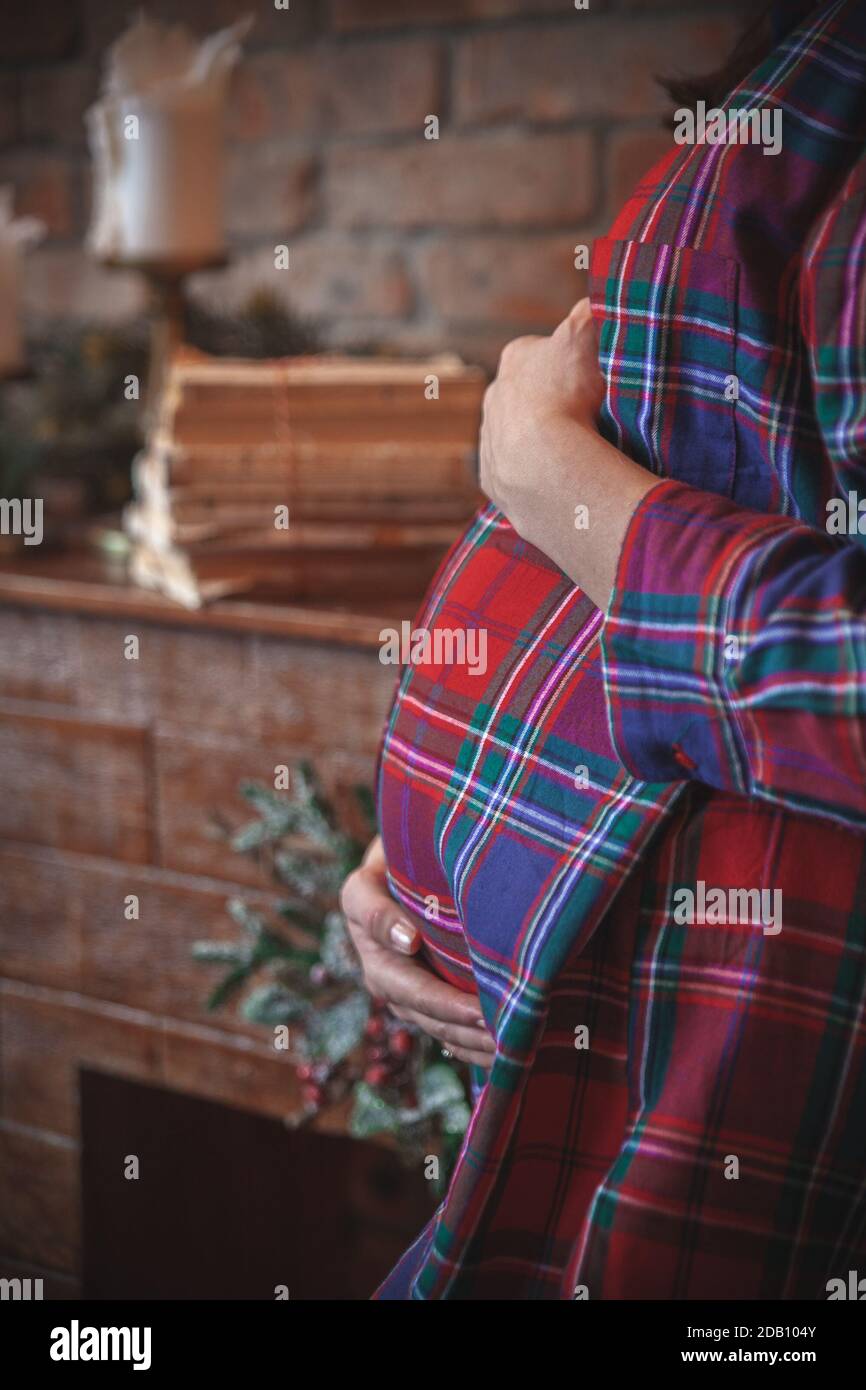 Christmas pregnancy. Pregnant woman holding her belly next to a cozy brick fireplace in a checkered red shirt. Cozy, rustic Christmas mood. Stock Photo