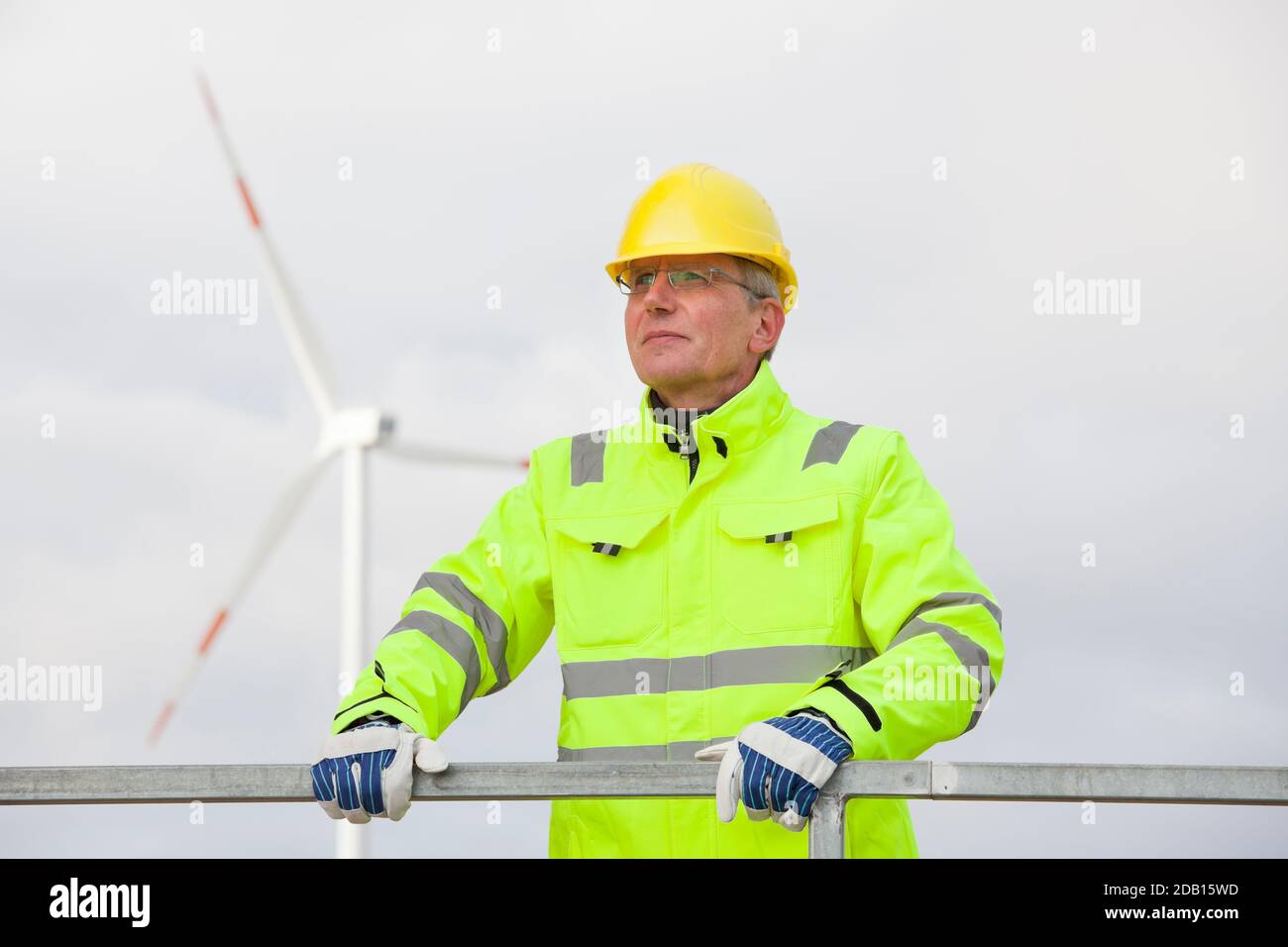 Mature engineer with hard hat and protective clothing in front of blurred wind turbine in the background Stock Photo